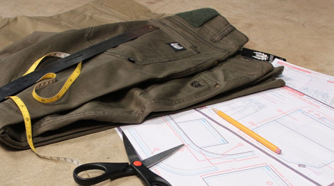 ruler, measuring tape and pants laid next to scissors and a pencil on top of design plans